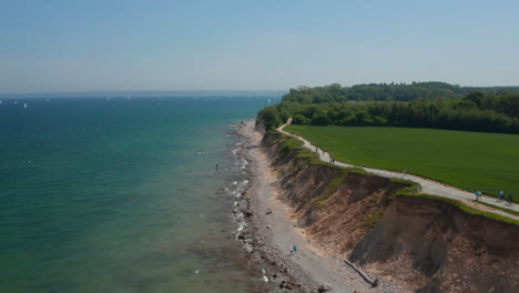 Aerial-view-of-German-coastline-facing-Baltic-sea,-vast-green-field-by-coast-with-pathway-people-passing-by,-clear-sky-day,-forward